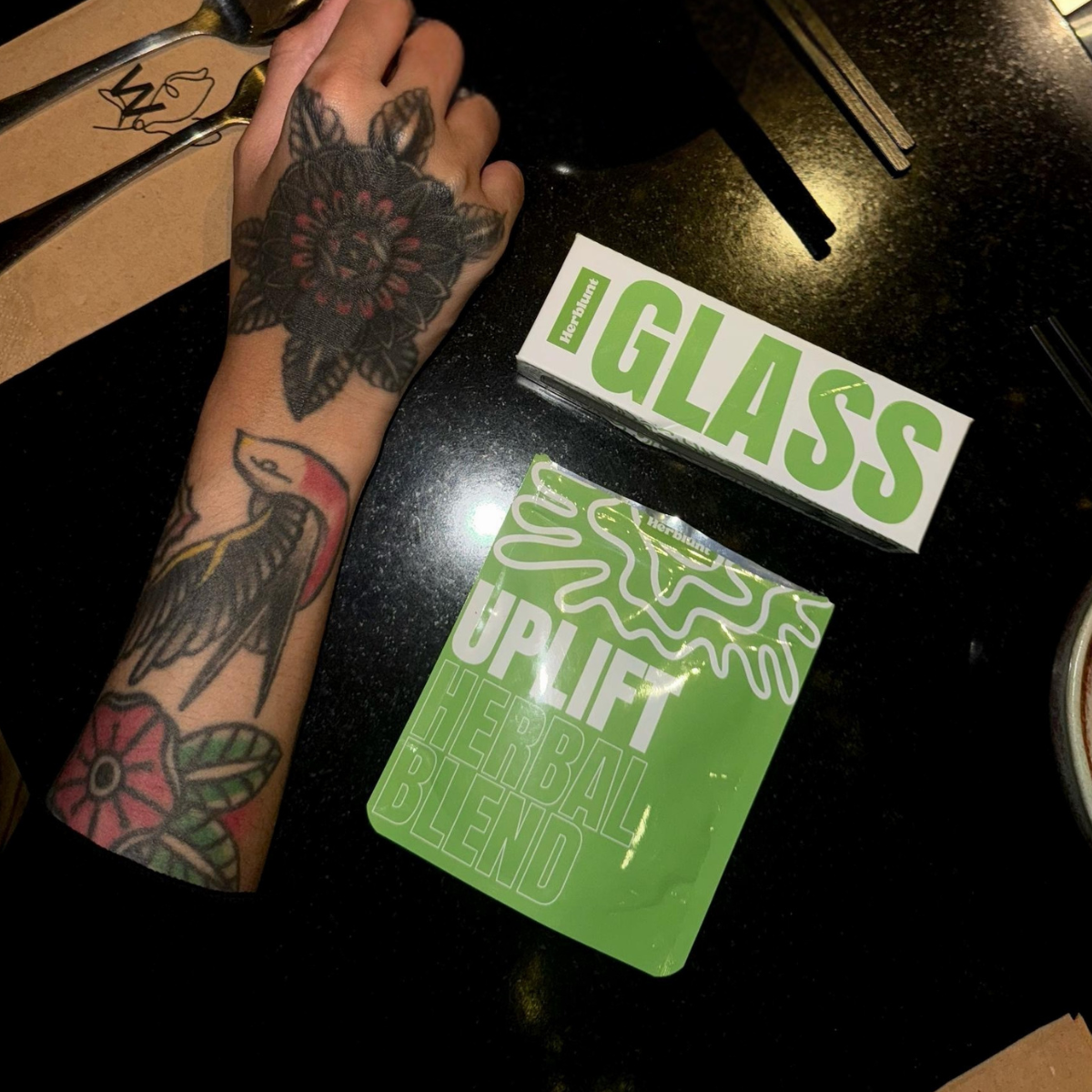 Tattooed arm with Herblunt 'Glass' and 'Uplift' products on a dark surface