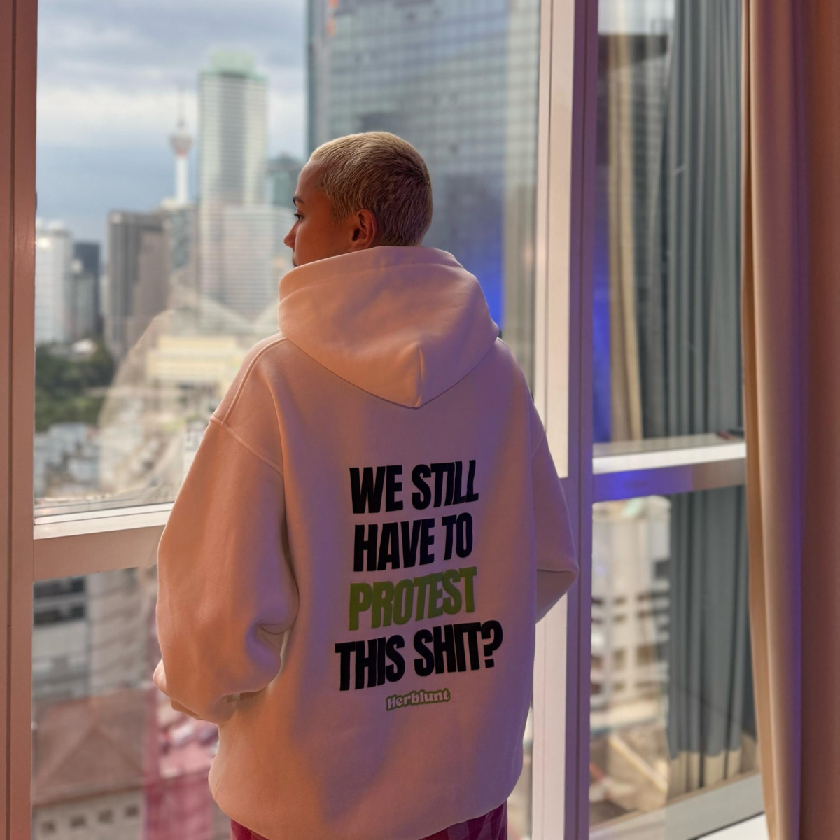 Person wearing a herblunt hoodie with text on the back, standing in front of a cityscape view.